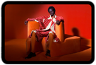 Vista frontal de un iPad mini que muestra una imagen nítida de una persona con un traje naranja sentada en una silla naranja en una habitación naranja
