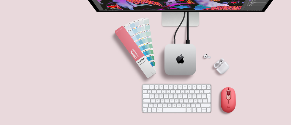 Mac mini, parte superior, aluminio color plata, esquinas redondeadas, logo de Apple en el centro en color negro brillante, conectada con un cable, monitor Studio Display parcialmente visible en la parte superior, AirPods 4, abiertos, muestra de una paleta de colores Pantone, teclado numérico y mouse