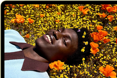 MacBook Neo, color amarillo cítrico, la pantalla muestra a una persona sonriente acostada en un campo de flores bajo el sol
