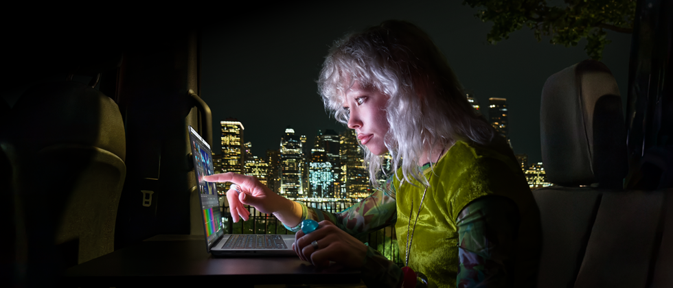 Una persona sentada en el asiento trasero de una van, la puerta del pasajero abierta muestra el horizonte de una ciudad de noche, MacBook Pro con pantalla PROLED XDR sobre un escritorio, la persona es iluminada por la pantalla mientras usa la funcionalidad Touch para editar un video en DaVinci Resolve Studio