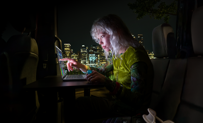 Una persona sentada en el asiento trasero de una van, la puerta del pasajero abierta muestra el horizonte de una ciudad de noche, MacBook Pro con pantalla PROLED XDR sobre un escritorio, la persona es iluminada por la pantalla mientras usa la funcionalidad Touch para editar un video en DaVinci Resolve Studio