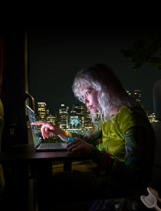 Una persona sentada en el asiento trasero de una van, la puerta del pasajero abierta muestra el horizonte de una ciudad de noche, MacBook Pro con pantalla PROLED XDR sobre un escritorio, la persona es iluminada por la pantalla mientras usa la funcionalidad Touch para editar un video en DaVinci Resolve Studio