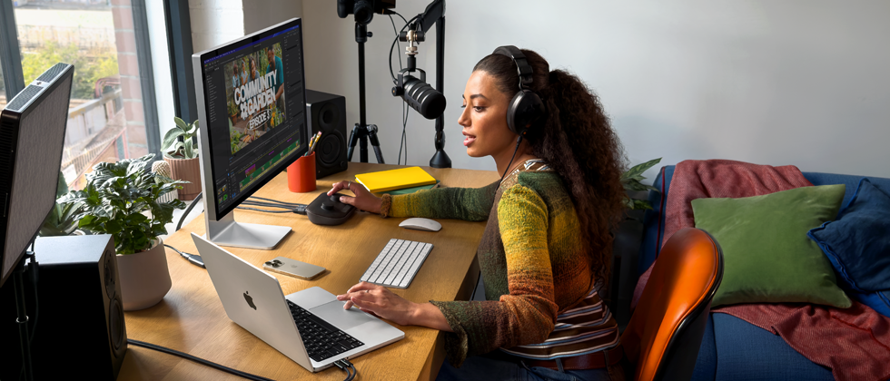 Una mujer en su casa trabajando desde su escritorio con una MacBook Pro conectada a dos monitores externos, usando audífonos y hablando en un micrófono