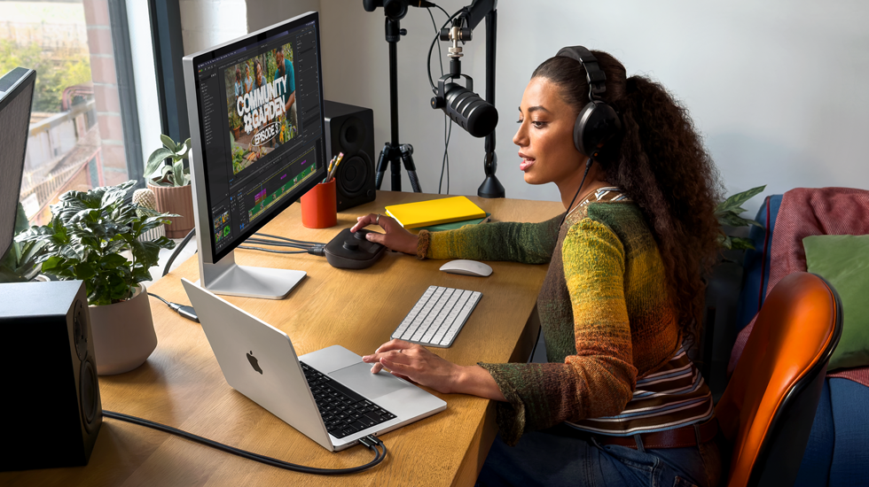 Una mujer en su casa trabajando desde su escritorio con una MacBook Pro conectada a un monitor externo, usando audífonos y hablando en un micrófono