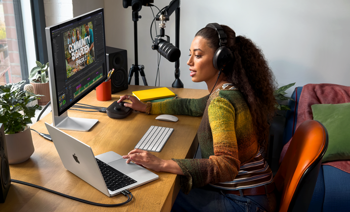 Una mujer en su casa trabajando desde su escritorio con una MacBook Pro conectada a un monitor externo, usando audífonos y hablando en un micrófono