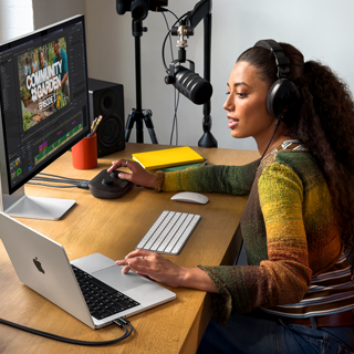 Una mujer en su casa trabajando desde su escritorio con una MacBook Pro conectada a un monitor externo, usando audífonos y hablando en un micrófono